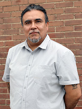 Upper torso portrait of Herbie, a man wearing a short sleeved button down shirt in front of a red brick wall