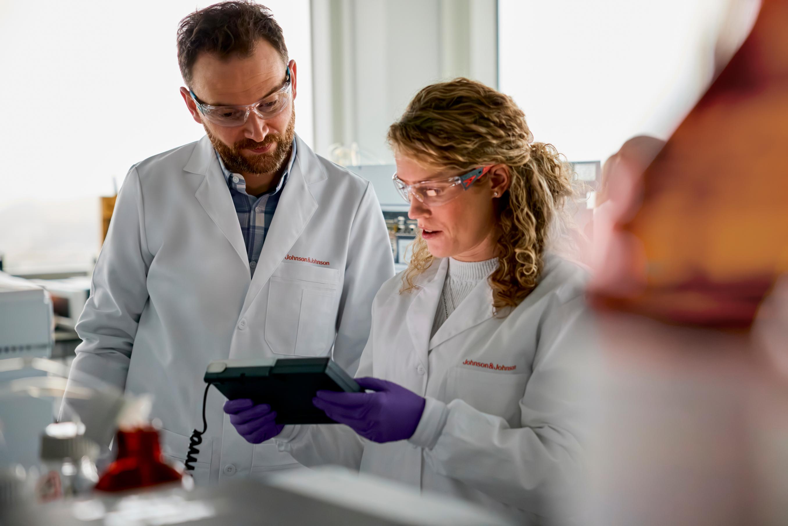 A man and woman in white lab coats look at a tablet