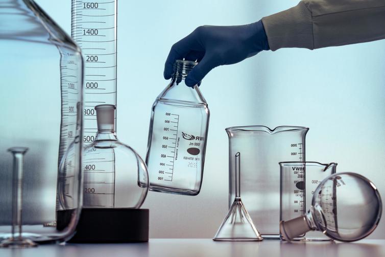 Gloved hand picking up flask with beakers and other lab instruments on table