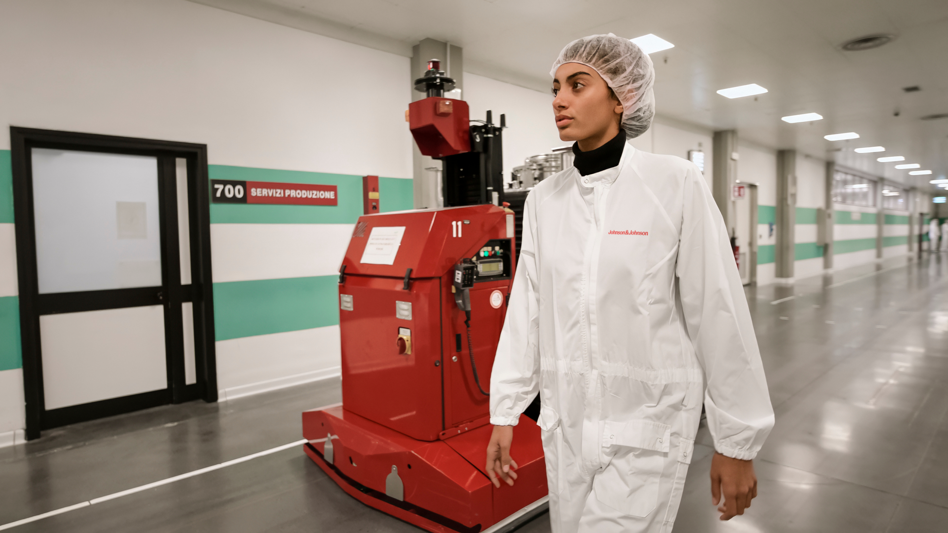 A clean, brightly lit corridor inside a pharmaceutical facility, where two operators in full cleanroom attire walk alongside an LGV (Laser Guided Vehicle).