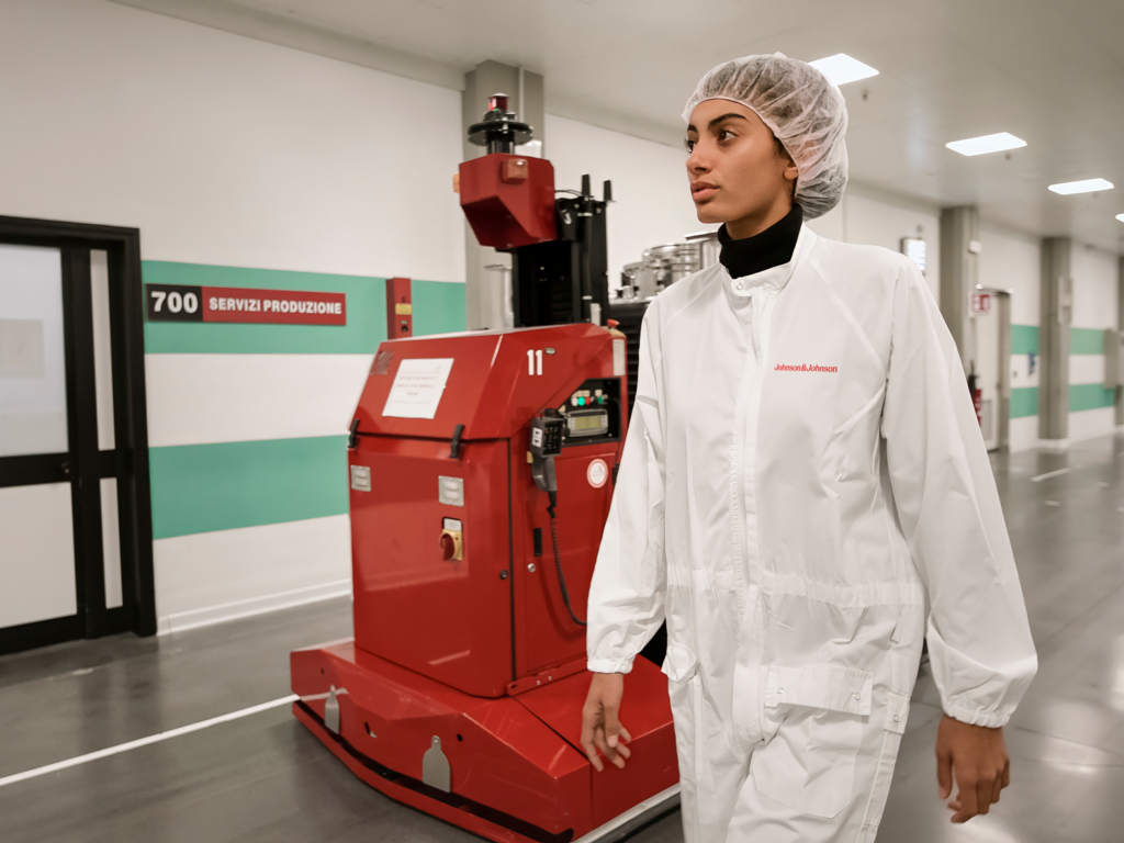 A clean, brightly lit corridor inside a pharmaceutical facility, where two operators in full cleanroom attire walk alongside an LGV (Laser Guided Vehicle).