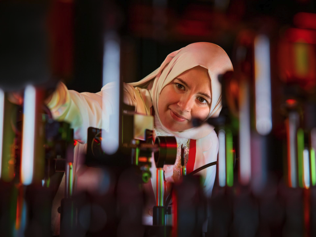 An optical R&D scientist adjusting components within a complex illuminated optical setup on a laboratory table.