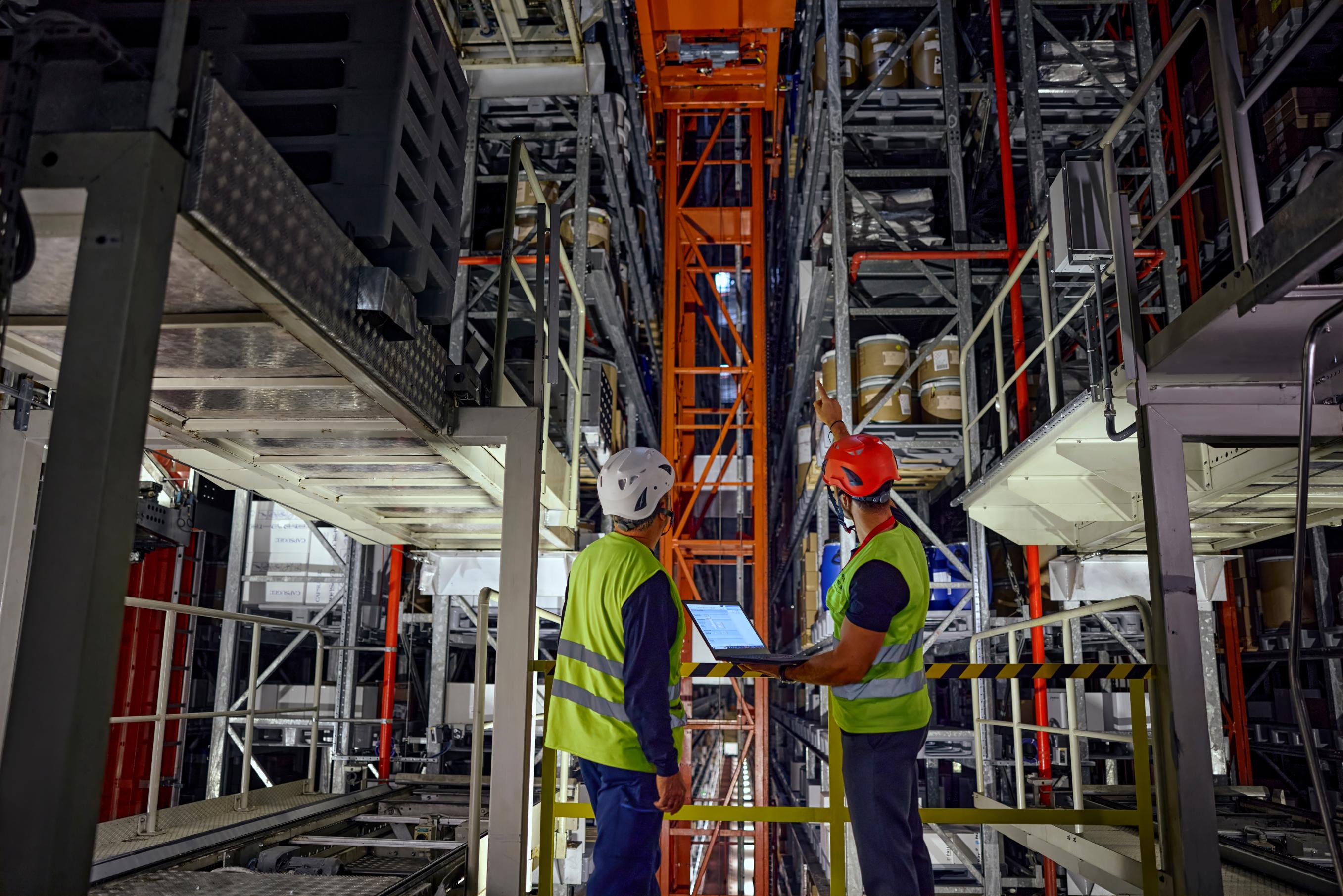 In the foreground, a warehouse manager and a specialist are inspecting the functionality of an automated crane system responsible for picking and placing materials within the storage racks
