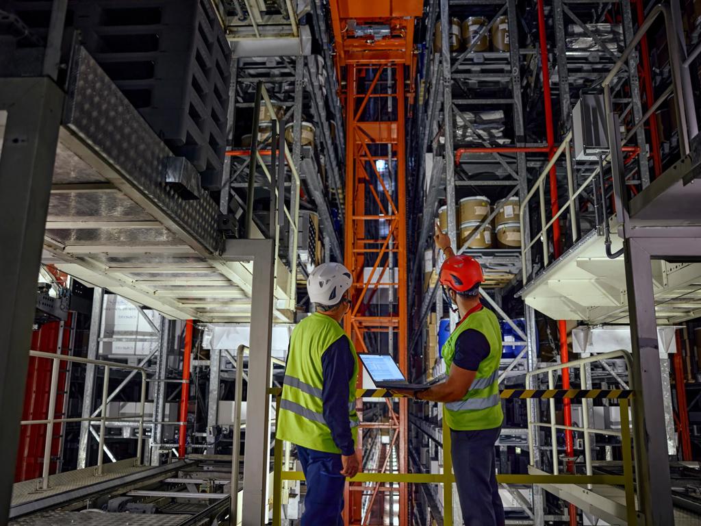 In the foreground, a warehouse manager and a specialist are inspecting the functionality of an automated crane system responsible for picking and placing materials within the storage racks