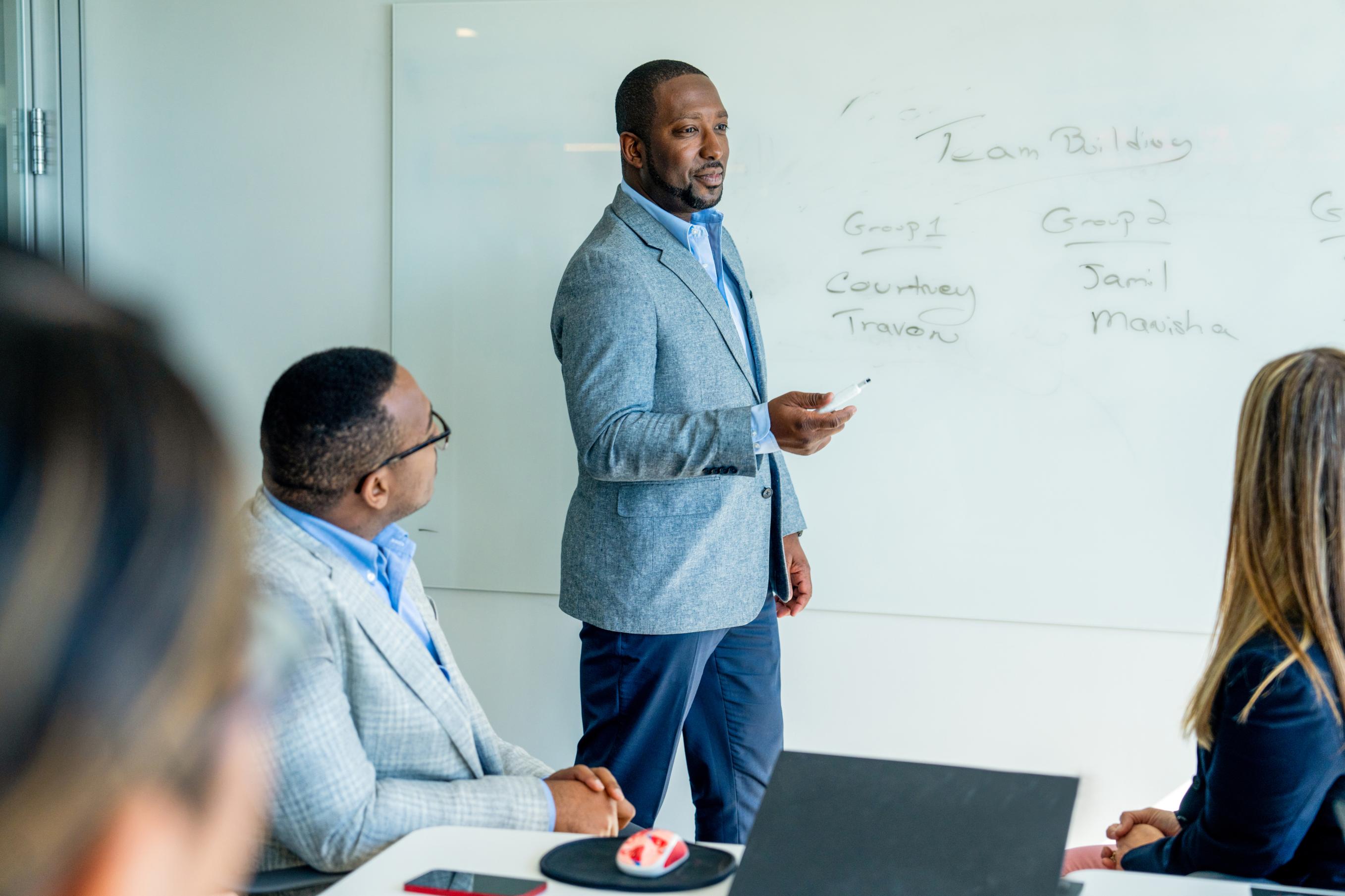 A man stands at a white board in an office leading a meeting