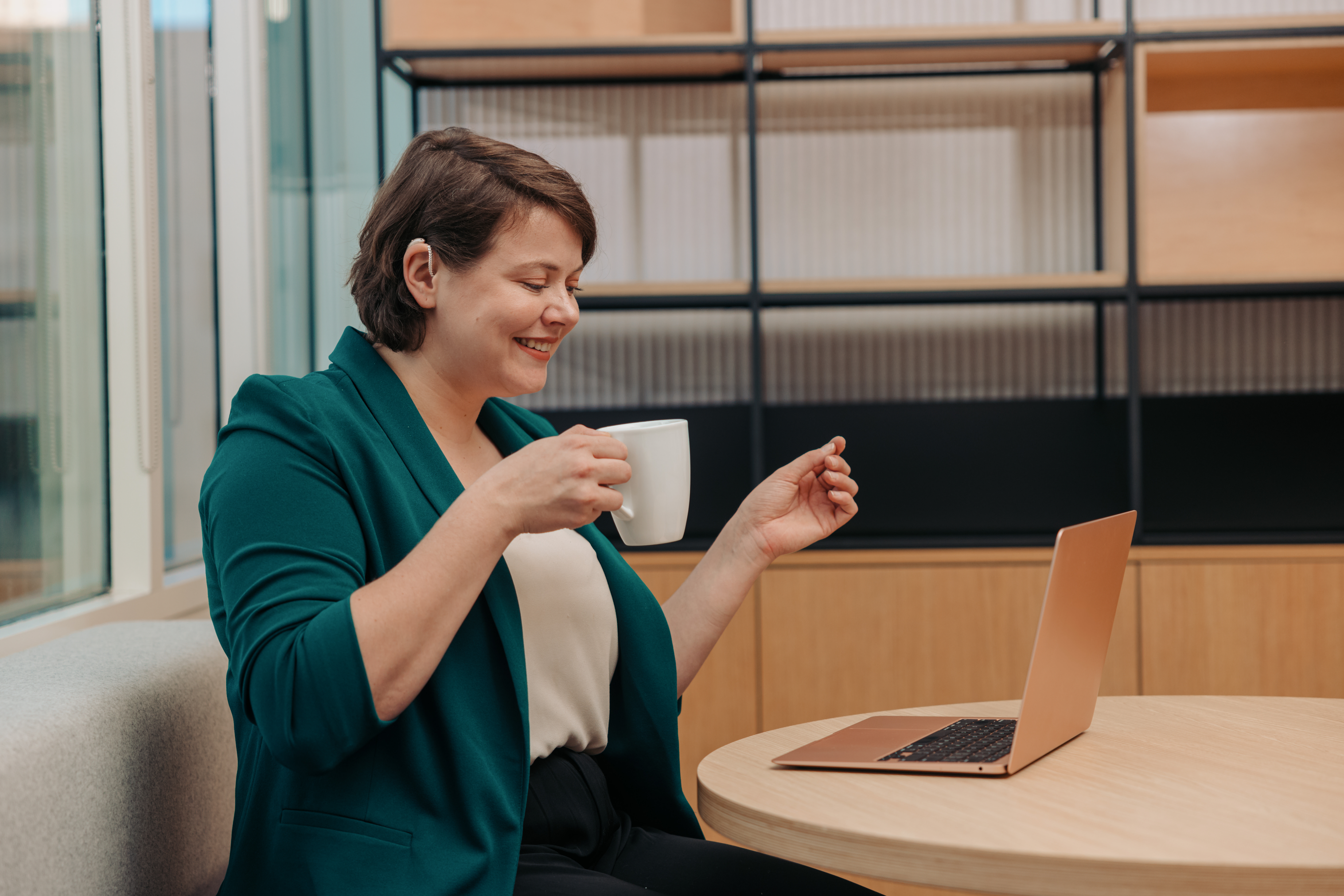 A woman works on her laptop while holding a cup of coffee in an office. Her hair is tucked behind her ear to show off their hearing aid.