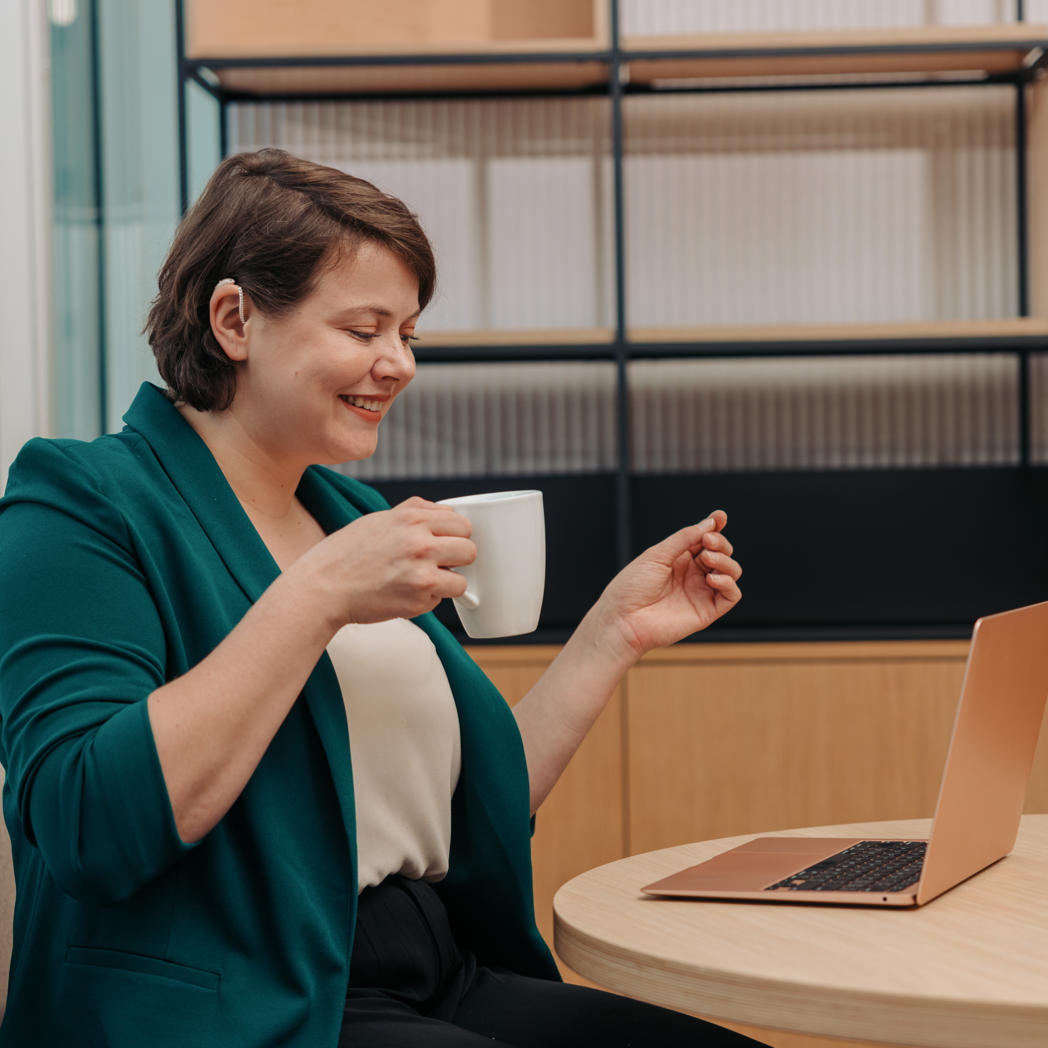 A woman works on her laptop while holding a cup of coffee in an office. Her hair is tucked behind her ear to show off their hearing aid.