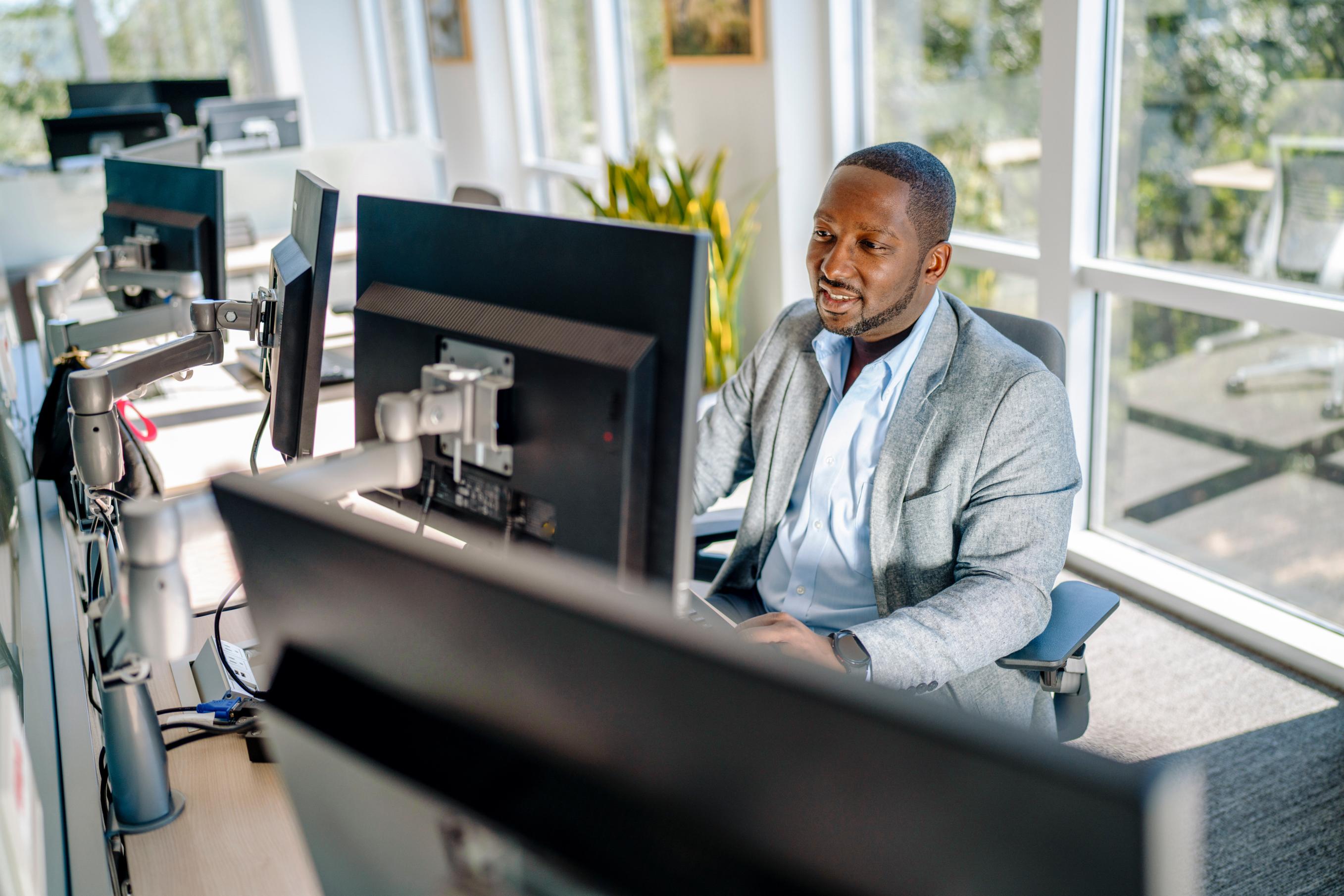 A man sits at a desk in front of windows working on a dual monitor computer