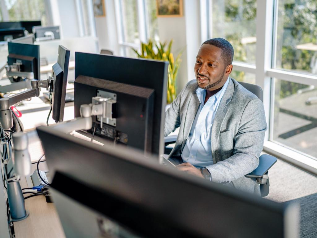 A man sits at a desk in front of windows working on a dual monitor computer